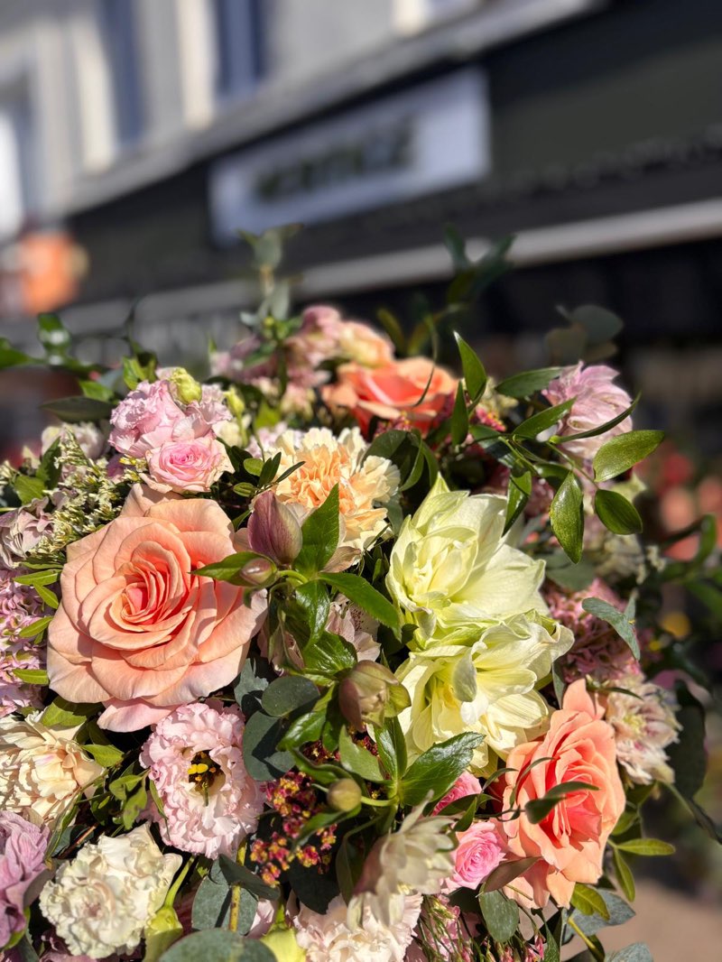 Bouquet pastel de roses pêche, œillets et amaryllis crème devant la devanture de Vertige